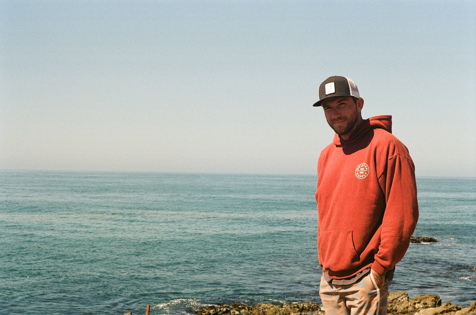 Charles in an orange hoodie and cap, standing by the ocean