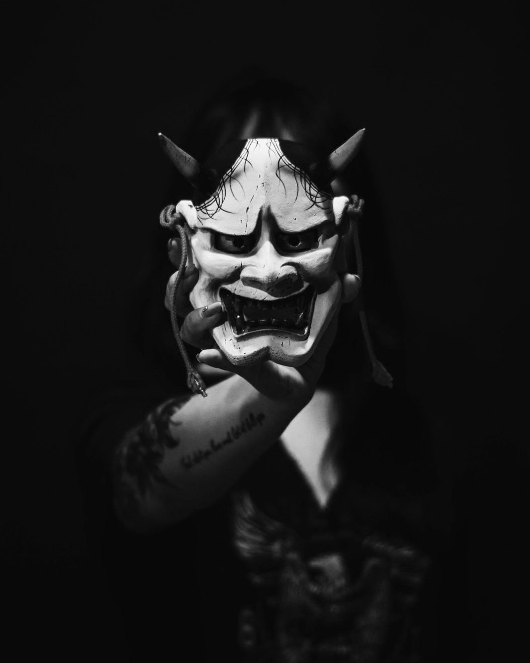 Black-and-white photo of a tattooed hand holding up a carved Hannya mask in the dark